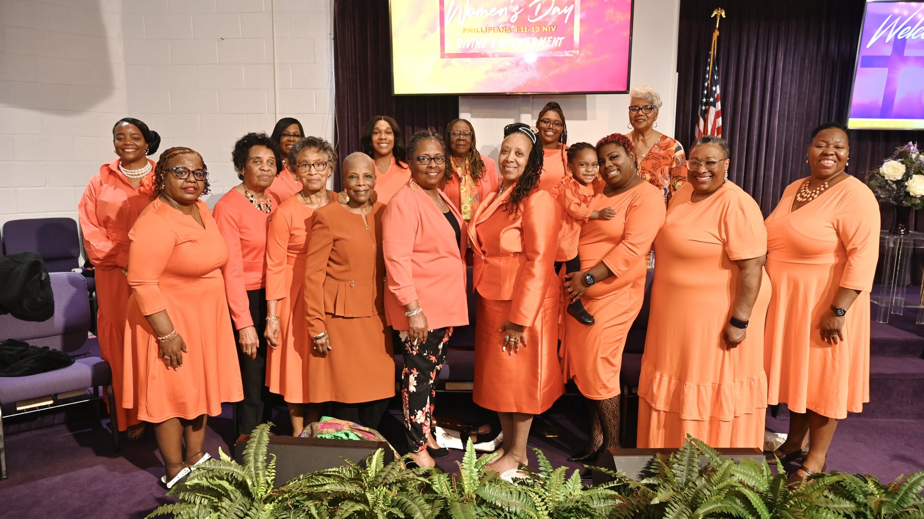 A group photo from Women's Day 2026: women of the church, dressed in coral and orange, gathered at the front of the sanctuary.